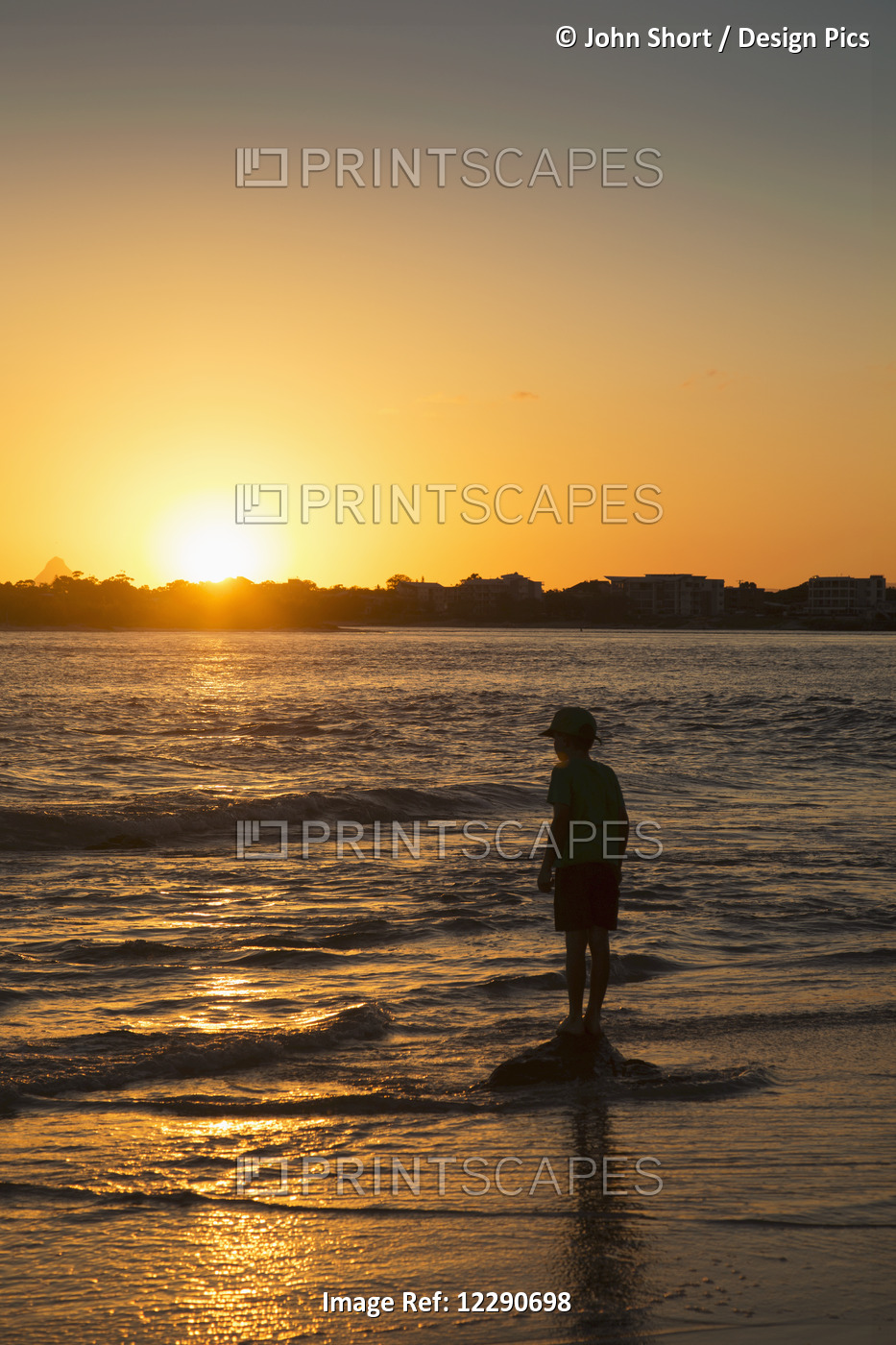 Silhouette Of A Young Boy Standing At The Water's Edge At Sunset; Caloundra, ...